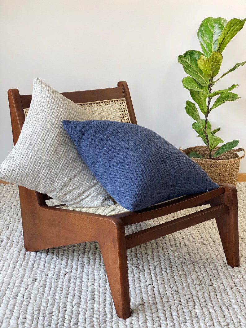 May include: A wooden chair with a woven cane seat and back, featuring two cushions. One cushion is light grey and the other is dark blue. The chair is sitting on a white fluffy rug.