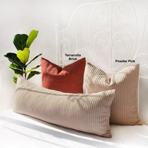 May include: Three decorative throw pillows on a white bed frame. The pillows are made of a textured fabric. The pillow on the left is a long bolster pillow in a light beige color. The middle pillow is a square pillow in a terracotta brick color. The pillow on the right is a square pillow in a powder pink color.