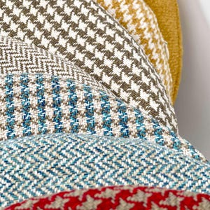 Puede incluir: Primer plano de una pila de telas estampadas en varios colores. Las telas incluyen pata de gallo, espiga y otros dise&ntilde;os tejidos en tonos marrones, azules, blancos y rojos. Adecuado para tapicer&iacute;a o decoraci&oacute;n del hogar.