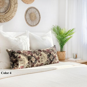 May include: A long, rectangular cushion with a geometric pattern in burgundy, beige, and green sits on a white bed. The cushion is labelled "Color 4". Woven wall hangings and a potted plant are in the background.