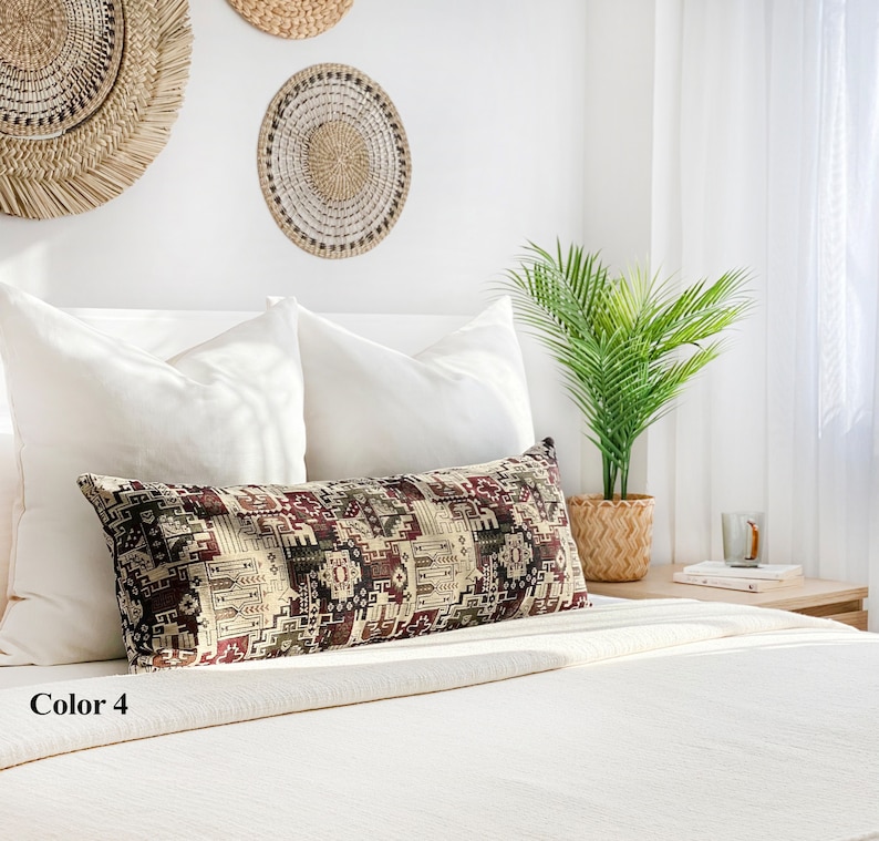 May include: A long, rectangular cushion with a geometric pattern in burgundy, beige, and green sits on a white bed. The cushion is labelled "Color 4". Woven wall hangings and a potted plant are in the background.