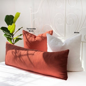 May include: Two decorative pillows, one in terracotta brick and one in white, are arranged on a bed with a white headboard. The pillows are made of a textured fabric with a cable knit design. The text "Terracotta Brick" and "White" is visible above the pillows.