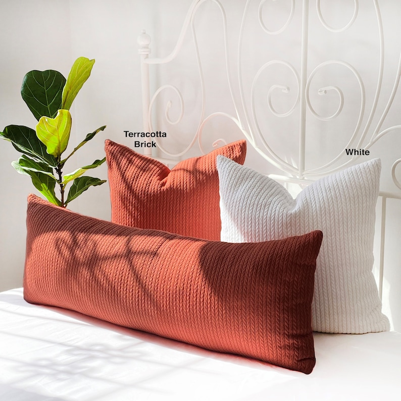 May include: Two decorative pillows, one in terracotta brick and one in white, are arranged on a bed with a white headboard. The pillows are made of a textured fabric with a cable knit design. The text "Terracotta Brick" and "White" is visible above the pillows.