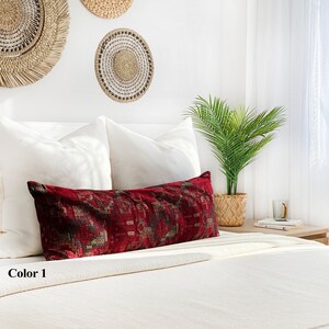 May include: A long, rectangular cushion with a deep red background and a patterned design in shades of brown and gold. The cushion is placed on a white bed with white pillows and decorative wall hangings in the background. The text "Color 1" is visible.