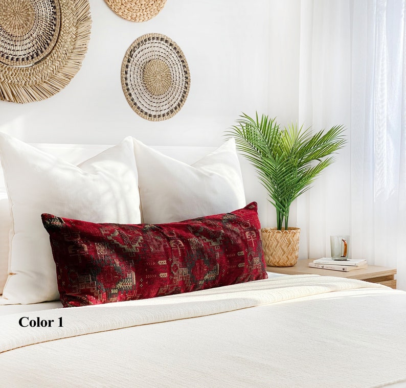 May include: A long, rectangular cushion with a deep red background and a patterned design in shades of brown and gold. The cushion is placed on a white bed with white pillows and decorative wall hangings in the background. The text "Color 1" is visible.