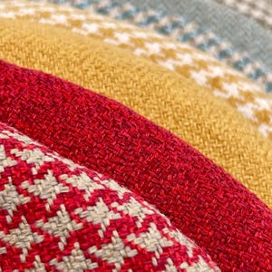 Puede incluir: Primer plano de varios textiles plegados en varios colores. Los textiles incluyen telas rojas, amarillas y verde azulado. Un textil tiene un estampado de pata de gallo rojo y blanco, mientras que otros tienen colores lisos o un estampado blanco.