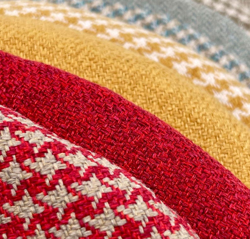 Puede incluir: Primer plano de varios textiles plegados en varios colores. Los textiles incluyen telas rojas, amarillas y verde azulado. Un textil tiene un estampado de pata de gallo rojo y blanco, mientras que otros tienen colores lisos o un estampado blanco.