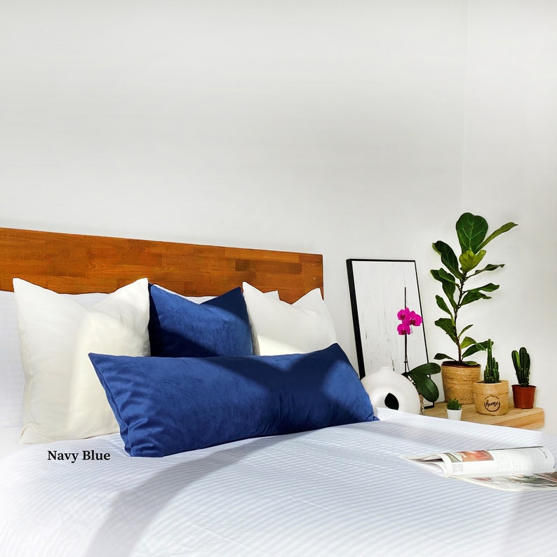 May include: A bed with white linens and a navy blue lumbar pillow. The bed has a wooden headboard and there are two white pillows and a navy blue square pillow on the bed. The bed is in a bedroom with a white wall and a plant in a pot on a side table.