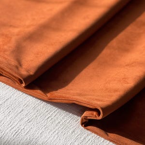 Peut inclure: Gros plan sur un tissu orange br&ucirc;l&eacute; pli&eacute;, &agrave; la texture douce et velout&eacute;e. Le tissu est drap&eacute;, r&eacute;v&eacute;lant les plis et les ombres. Le mat&eacute;riau semble &ecirc;tre un textile moelleux, peut-&ecirc;tre pour la tapisserie ou l'habillement.