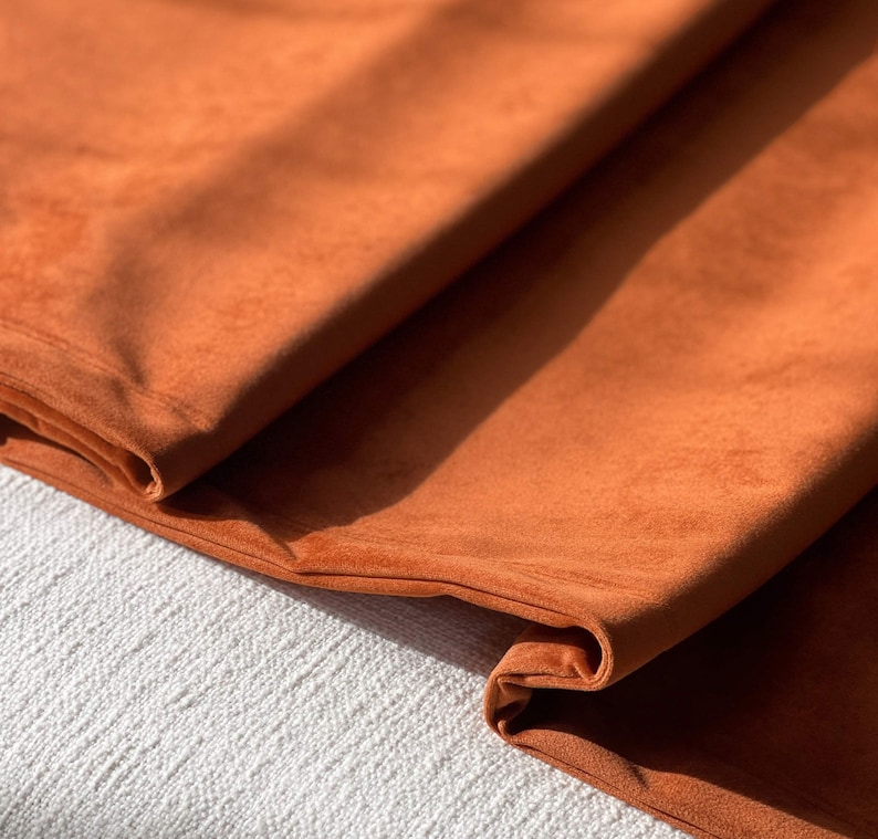 Peut inclure: Gros plan sur un tissu orange br&ucirc;l&eacute; pli&eacute;, &agrave; la texture douce et velout&eacute;e. Le tissu est drap&eacute;, r&eacute;v&eacute;lant les plis et les ombres. Le mat&eacute;riau semble &ecirc;tre un textile moelleux, peut-&ecirc;tre pour la tapisserie ou l'habillement.