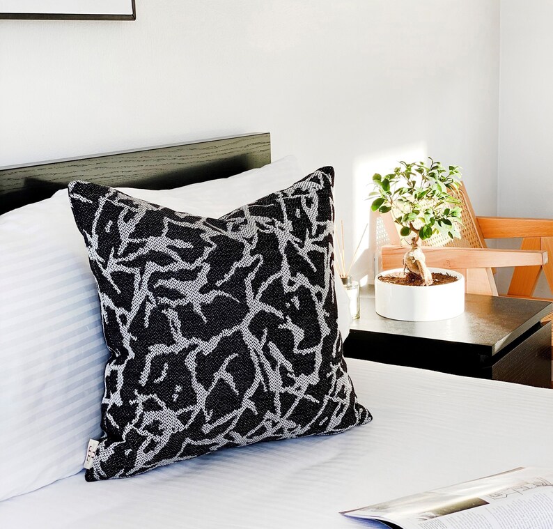 May include: A black and white patterned throw pillow with a geometric design. The pillow is on a white bed with a black headboard.