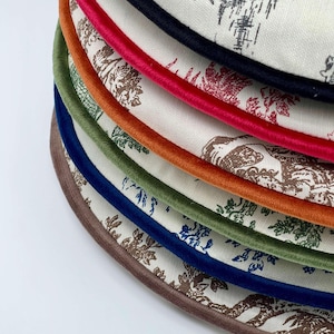 May include: A stack of round fabric coasters with a floral pattern in shades of brown and blue. Each coaster has a different colored trim, including red, orange, green, and blue. The coasters are made of a soft, textured fabric.