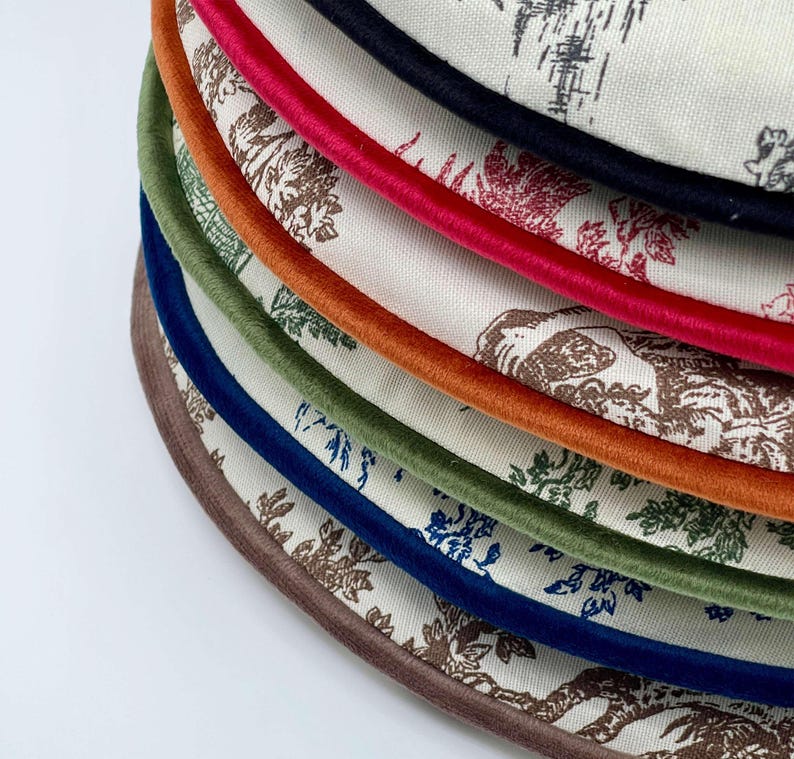 May include: A stack of round fabric coasters with a floral pattern in shades of brown and blue. Each coaster has a different colored trim, including red, orange, green, and blue. The coasters are made of a soft, textured fabric.