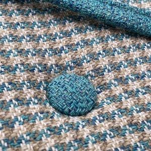 Puede incluir: Primer plano de una tela texturizada con un patr&oacute;n tejido en tonos turquesa, blanco y beige. Se ve un bot&oacute;n turquesa, que a&ntilde;ade un toque de color. La tela parece ser material de tapicer&iacute;a, posiblemente para muebles o decoraci&oacute;n del hogar.