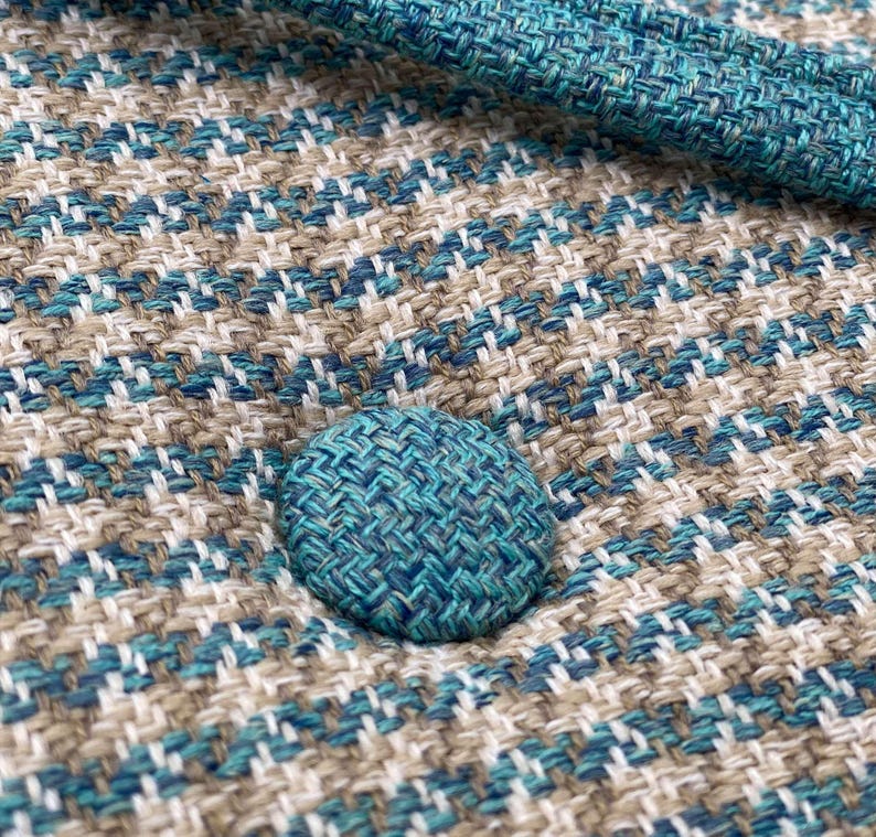 Puede incluir: Primer plano de una tela texturizada con un patr&oacute;n tejido en tonos turquesa, blanco y beige. Se ve un bot&oacute;n turquesa, que a&ntilde;ade un toque de color. La tela parece ser material de tapicer&iacute;a, posiblemente para muebles o decoraci&oacute;n del hogar.