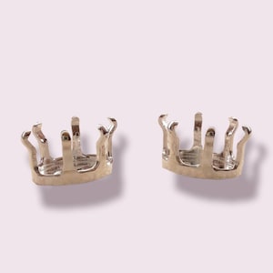 May include: Two silver crown-shaped settings for gemstones. The settings are open on the bottom and have a round opening in the center.