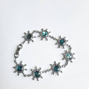 May include: A silver bracelet with a chain and seven round charms. Each charm is a silver ship's wheel with a blue and green abalone shell in the center.