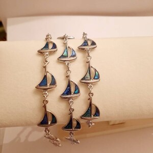 May include: Three silver bracelets with blue and green abalone shell sailboat charms. The bracelets are layered on a white surface.