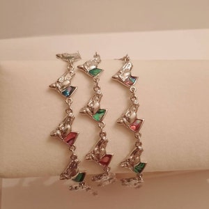 May include: Three silver bracelets with a geometric design. Each bracelet features a repeating pattern of small, triangular-shaped charms with iridescent green, pink, and blue accents.
