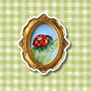 May include: An oval-shaped, ornate gold-colored frame containing a small painting of a red ladybug with black spots resting on a green leaf. The frame features intricate embossed detailing along its edges and a classic, decorative border design.