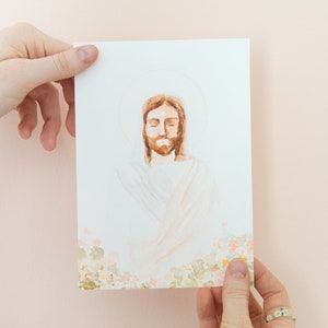May include: A watercolour illustration of a figure with a halo, eyes closed, and a beard. The artwork is on a white card with a floral design at the bottom. The card is held by two hands against a light pink background.