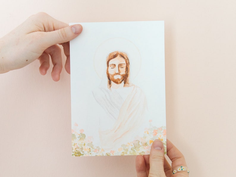 May include: A watercolour illustration of a figure with a halo, eyes closed, and a beard. The artwork is on a white card with a floral design at the bottom. The card is held by two hands against a light pink background.