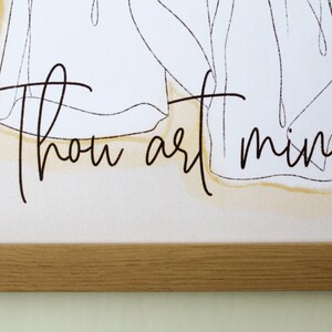 May include: A framed art print featuring the words "Thou art mine" in elegant black script. The artwork includes a minimalist line drawing of figures, set against a soft, neutral background with a subtle yellow wash. The frame is made of light-colored wood.
