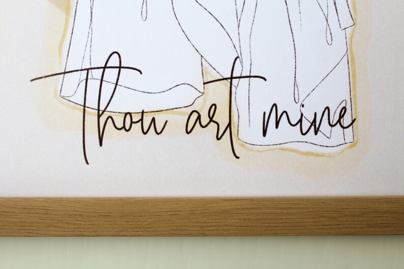 May include: A framed art print featuring the words "Thou art mine" in elegant black script. The artwork includes a minimalist line drawing of figures, set against a soft, neutral background with a subtle yellow wash. The frame is made of light-colored wood.