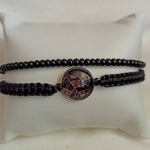 May include: A black beaded bracelet with a black cord and a round, silver-toned charm with a geometric design in shades of pink and brown.