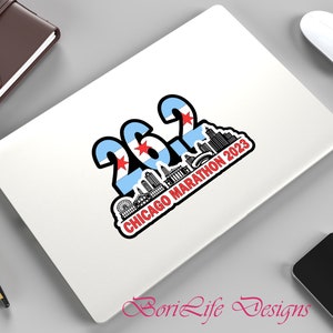 May include: A white laptop with a sticker that says "26.2 Chicago Marathon 2023" with a skyline of Chicago and the Chicago flag.