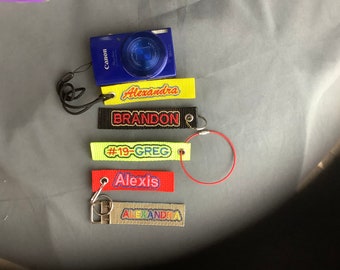 May include: A blue digital camera with a black strap and five colorful keychains with embroidered names: "Alexandra", "Brandon", "#19-Greg", "Alexis", and "Alexandra".