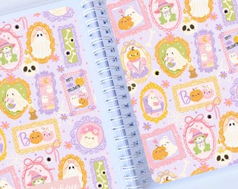 May include: Open planner with a spiral binding, featuring a light purple background filled with Halloween-themed illustrations. The designs include ghosts, pumpkins, and framed characters in pastel colors. The text "Happy Halloween" is visible.