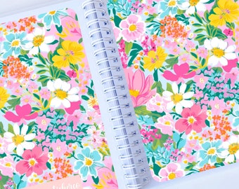 May include: A spiral bound notebook with a floral pattern on the cover. The cover features a variety of pink, yellow, blue, and green flowers on a light blue background. The text "Laurastickerei" is printed on the front cover.
