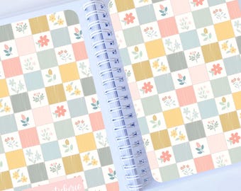 May include: A spiral bound notebook with a floral pattern on the cover. The cover is a checkered pattern of pink, yellow, blue, and gray squares with small floral designs in each square. The text "laurasstickers" is written in pink on the front cover.