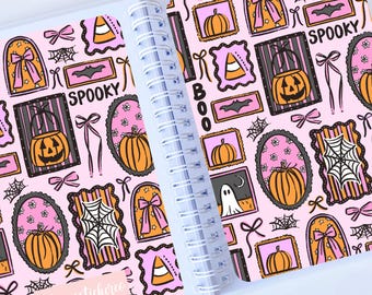 May include: Open notebook with a pink Halloween-themed design. The pattern includes pumpkins, ghosts, spiderwebs, and the word "Spooky." The notebook has a spiral binding and clear plastic covers.