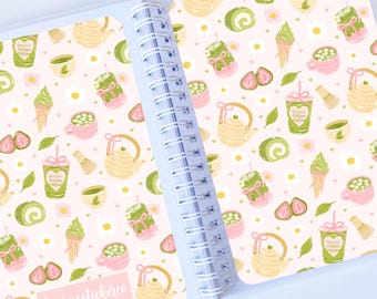 May include: Open notebook with a spiral binding, featuring a repeating pattern of matcha tea elements. The design includes teapots, cups, ice cream, strawberries, and matcha drinks in shades of green, pink, and yellow on a light pink background. The text "laurasstickerei" is visible.