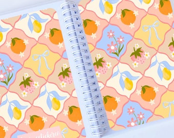 May include: A spiral bound notebook with a colorful pattern of lemons, oranges, strawberries, and flowers on a light blue, pink, and yellow background. The cover has the text "LaurasStickers" on the bottom right corner.