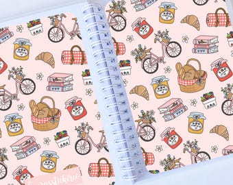 May include: A pink and white spiral bound notebook with a whimsical pattern of bicycles, jars of jam, croissants, and picnic baskets. The text "laurasticklebee" is printed on the cover.