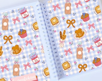 May include: Open notebook with a spiral binding, featuring a blue and white checkered pattern. The pattern includes illustrations of bread, pancakes, cupcakes, bows, and jam jars. A pink label with text is visible.