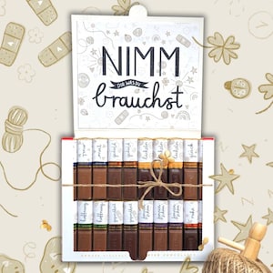 Könnte beinhalten: Eine weiße Geschenkbox mit Schokoladenriegeln, die mit Bindfaden zusammengebunden sind. Der Deckel trägt den deutschen Schriftzug "Nimm dir was du brauchst". Die Schokoriegel sind in verschiedenen Verpackungen. Geschenkthema.