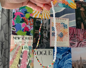 May include: Two necklaces, one with white pearl beads and the other with colourful beads. The necklaces are held in a hand against a background of colourful magazine covers.