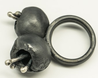 May include: A unique silver ring featuring three dark gray, textured, organic-shaped elements, each with silver accents. The ring band is a simple, dark gray band. The design is modern and artistic.