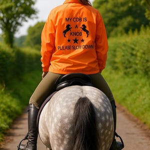 May include: A person riding a grey horse, wearing an orange riding jacket with the text "MY COB IS A KNOB PLEASE SLOW DOWN". The rider wears a black helmet and olive-green riding trousers. The horse is on a country lane.