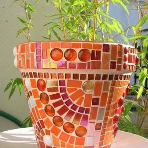 May include: A mosaic-tiled flower pot in shades of orange, red, and pink. The pot is decorated with small, irregularly shaped tiles and larger, round, glass-like accents. Green bamboo plants are visible in the background.