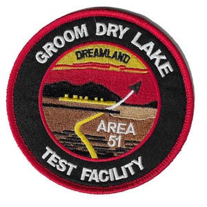 Op de afbeelding: Een geborduurde patch in rood, wit en zwart met de tekst "Groom Dry Lake Test Facility" en een kaart van "Area 51" met de tekst "Dreamland" in een rode cirkel.