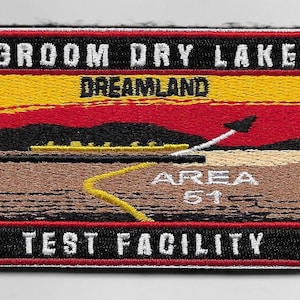 Puede incluir: Un parche bordado en blanco y negro con el texto "Groom Dry Lake Dreamland Area 51 Test Facility". El parche presenta una puesta de sol roja y amarilla con una silueta negra de un avión volando sobre un paisaje desértico.