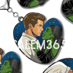 Puede incluir: Un llavero en forma de corazón con una ilustración de una pareja. La mujer tiene la piel verde y el hombre lleva una camisa blanca. El fondo es azul oscuro con estrellas y el texto "SALEM365" es visible. Tiene un cierre plateado.