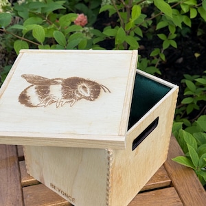 May include: A light-colored wooden box with a square lid featuring a laser-engraved bumblebee design. The side of the box displays the name BEYONCE and the number 8/80. The interior of the box is lined with dark green fabric and has a cutout handle.