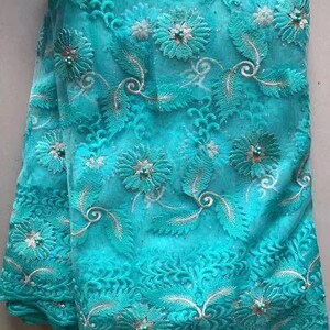 May include: A vibrant turquoise fabric with intricate floral embroidery. The design features detailed flowers, leaves, and swirling patterns, accented with small, pearl-like embellishments. The fabric has a delicate, textured appearance, suitable for apparel or decorative projects.