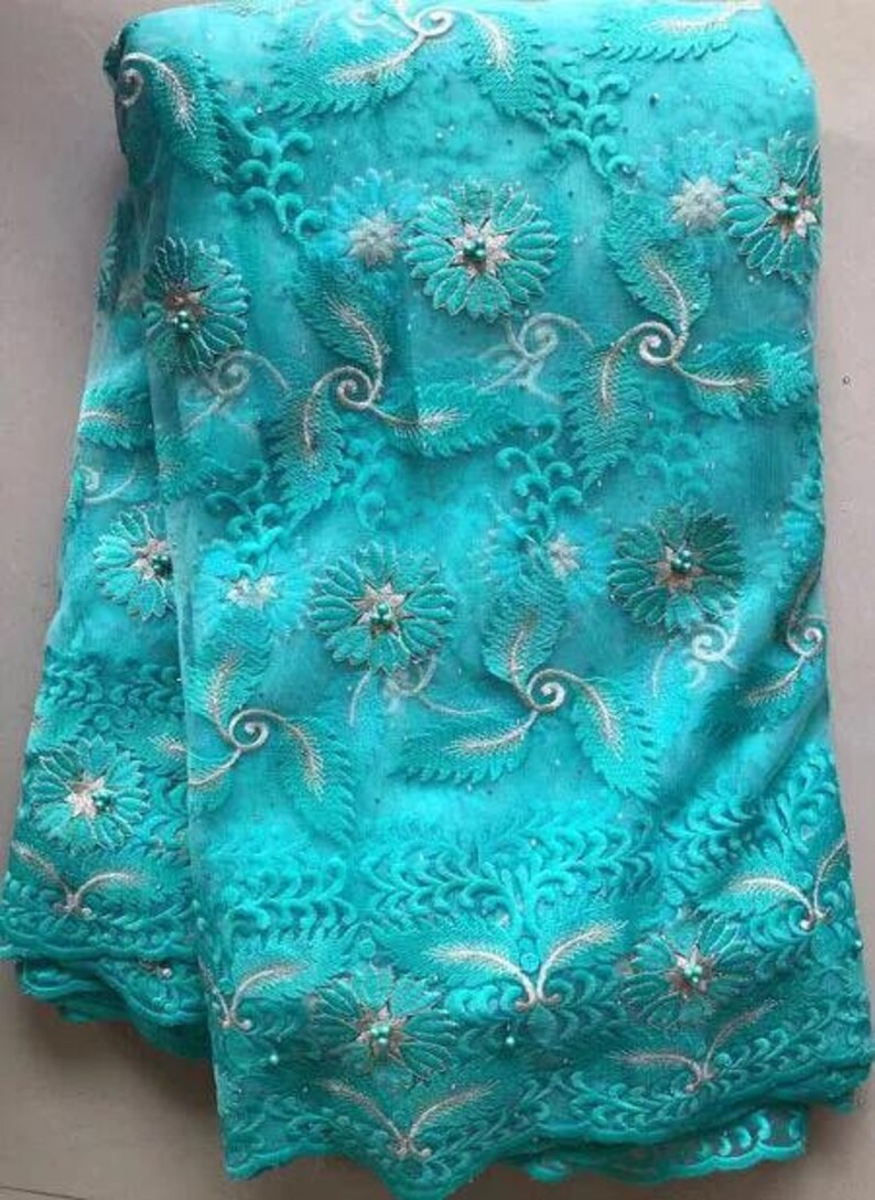 May include: A vibrant turquoise fabric with intricate floral embroidery. The design features detailed flowers, leaves, and swirling patterns, accented with small, pearl-like embellishments. The fabric has a delicate, textured appearance, suitable for apparel or decorative projects.